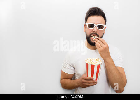 Photo de jeune homme debout et à la recherche sur l'appareil-photo. Sa bouche est pleine de pop-corn. Il est titulaire d'petit seau avec Crunchy Corn et continuer à manger. Isolé sur fond blanc. Banque D'Images