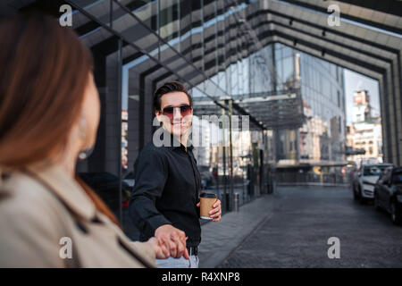 Cheerful young homme debout à l'extérieur avec femme et tenir sa main. Il la regarde et sourire. Guy détient tasse de boisson et sourire. Elle le regarde. Banque D'Images