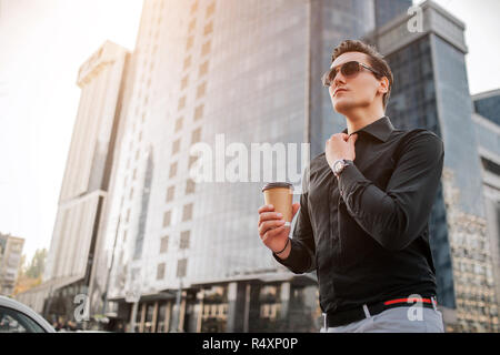 Les jeunes grave mn détient la main au collet de chemise et attendons avec intérêt. Il a tasse de boisson. Guy se place en avant des gratte-ciel. Banque D'Images