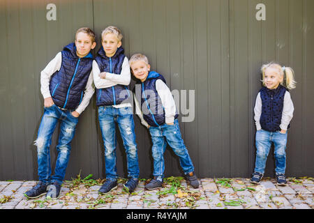 Funny Portrait grande famille de trois frères et sœur posant debout sur fond de croissance de mur en pleine croissance. Tout aussi élégamment vêtus de gilets et bleu jeans. Thème fille en cercle masculin Banque D'Images