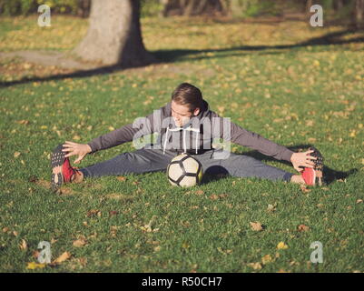 Joueur de football qui s'étend avec le football dans le parc dans le cadre d'une journée ensoleillée d'automne Banque D'Images
