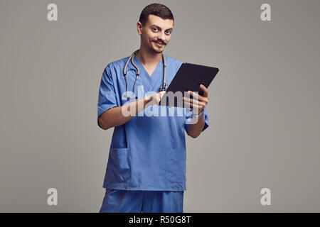 Portrait de jeune médecin arabe confiant avec moustache fantaisie en bleu est comprimé sur fond gris studio Banque D'Images