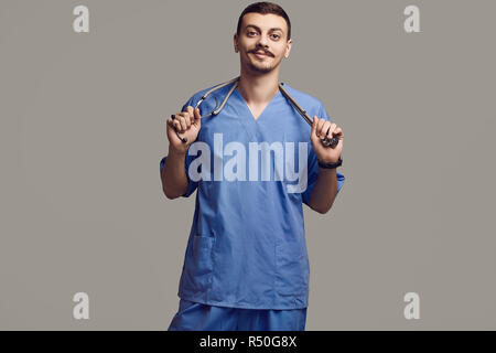 Portrait de jeune médecin arabe confiant avec moustache fantaisie en bleu sur fond gris studio Banque D'Images