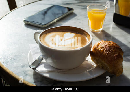 Petit-déjeuner avec l'art du latte sur une tasse de cappuccino avec un smart-phone et un jus d'orange sur une table. Banque D'Images