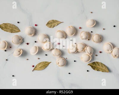 Schéma des pelmeni russes non cuits congelés avec des grains de poivre et les feuilles de laurier sur table en marbre blanc. Mise en page créative de boulettes. Belles matières dispersées quenelles. Vue de dessus ou de télévision. Banque D'Images