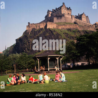 Années 1960, historique, un groupe de femmes siégeant ensemble sur l'herbe dans les jardins de Princes Street au-dessous de l'impressionnante forteresse de Château d'Edimbourg qui domine l'horizon, d'Édimbourg, en Écosse. Le château est situé sur Castle Rock, un énorme rocher volcanique formé il y a plusieurs millions d'années. Banque D'Images