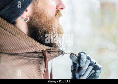 Portrait d'hiver jeune homme de profil. Main gantée zips veste une barbe sous le menton couvert de glaçons et gouttelettes congelées. Concept Gel Banque D'Images