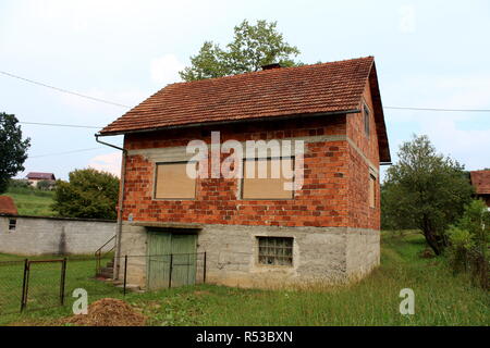 Red Brick abandonné inachevé maison de famille avec fenêtre fermée stores et portes de garage en bois délabrée entourée d'herbe non coupée et clear sky Banque D'Images