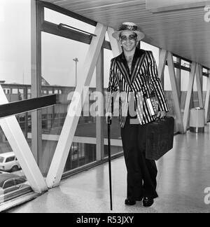 Pop star Elton John portant un chapeau garni de fleurs et de plumes, à l'arrivée à l'aéroport de Heathrow à partir de Los Angeles, où il a été l'enregistrement d'un nouvel album. Il est de retour pour un séjour de trois semaines et le haut de sa liste de ses priorités est l'demain match de football à Huddersfield où Elton sera criant pour son équipe préférée.. Watford. 30 août 1974. Banque D'Images