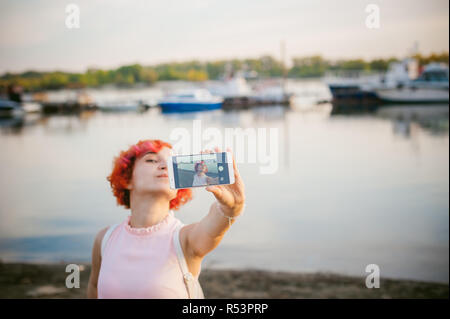 Fille en robe rose pâle aux cheveux rouges et son sac à dos marchant le long de rivière, photos d'eux-mêmes sur leur mobile téléphone appareil photo, contre fond de bateaux amarrés sur une chaude journée d'été Banque D'Images