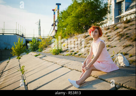 Fille en robe rose pâle aux cheveux rouges et son sac à dos à marcher le long de la rivière, assis sur la rive de sable Banque D'Images