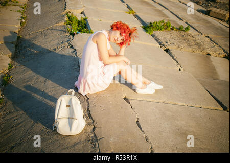 Fille en robe rose pâle aux cheveux rouges et son sac à dos à marcher le long de la rivière, assis sur la rive de sable Banque D'Images