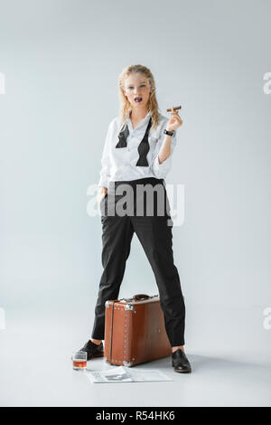 Belle fille à la mode de fumer un cigare en se tenant sur le gris avec sac de voyage vintage, presse et verre de whisky Banque D'Images