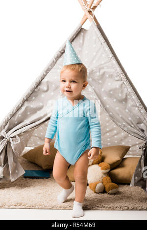 Bébé garçon dans party hat standing in front of wigwam avec oreillers et ours isolated on white Banque D'Images