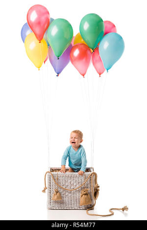 Bébé garçon pleurant et debout dans panier en osier avec des ballons multicolores isolated on white Banque D'Images