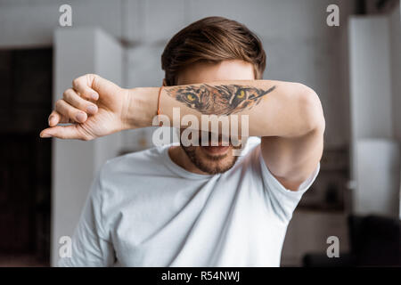 Beau jeune homme couvrant le visage avec le bras avec tigre tatoué à la maison Banque D'Images