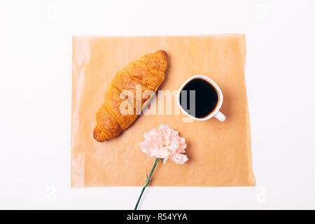 Mise à plat de la composition féminine petit-déjeuner simple de croissant et tasse de café noir avec fleur rose sur fond blanc, vue du dessus. Banque D'Images