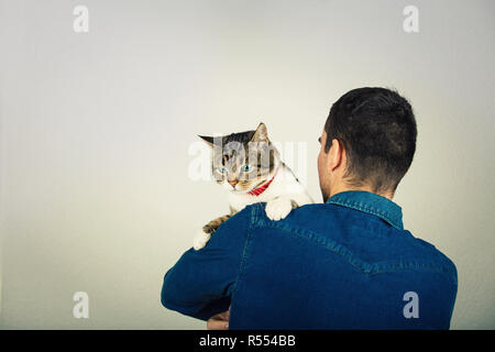 Vue arrière du jeune homme portant veste bleu jeans serrant un mignon et curieux cat holding à épaule. L'amour et du soin des animaux plus isolés grey wall background Banque D'Images