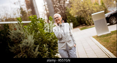 Smiling senior businesswoman parler au téléphone alors qu'elle est debout devant un immeuble de bureaux Banque D'Images