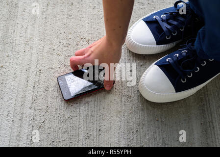 Femme jambes en formateurs près de broken fallen phone écran fissuré sur fond de béton. Téléphone cueillette à la main Banque D'Images