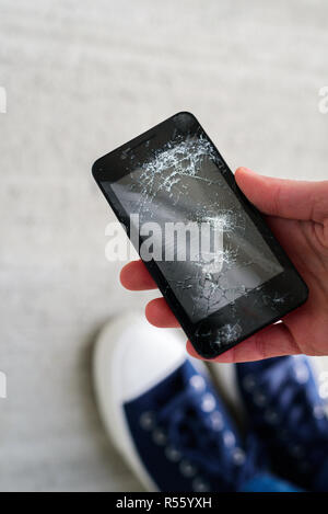 Femme jambes en formateurs près de broken fallen phone écran fissuré sur fond de béton. Téléphone cueillette à la main Banque D'Images