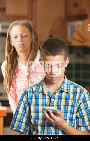 Mère tente d'oeil comme fils de l'adolescence vérifie son téléphone mobile Banque D'Images