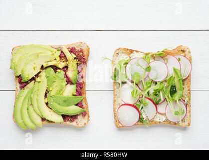 Deux toasts à l'avocat avec la purée d'avocat et de betterave, tranches d'avocat, radis, pousses de pois de neige et de fromage de chèvre sur fond de bois blanc Banque D'Images