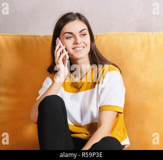 Jolie fille brune aux cheveux longs assis sur le canapé jaune à la lumière salon et chatter sur le smartphone avec son amie, la technologie moderne, communication concept Banque D'Images