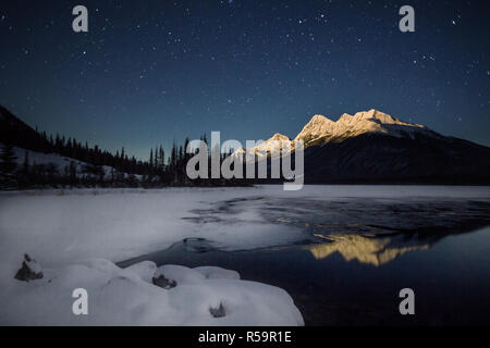 La haute montagne couverte de neige, dans la lumière de la pleine lune avec une demi lac gelé sous ciel plein d'étoiles, Parc national de Banff, Canada Banque D'Images