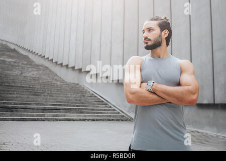 Photo de solides et bien construits young man standing et poser sur l'appareil-photo. Il regarde à gauche. L'homme conserve les mains croisées. Il a l'air mince et attrayant Banque D'Images