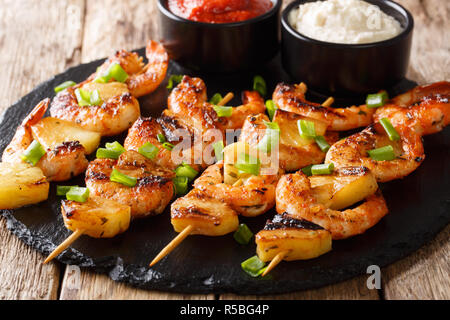 Brochettes de crevettes grillées à l'ananas frais et les oignons verts servis avec des sauces close-up sur une plaque en ardoise sur une table de bois horizontal. Banque D'Images