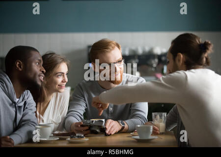 Jeune brun rouge afficher les images sur l'appareil photo à des amis, assis dans coffeeshop ensemble s'amuser, partager des souvenirs homme millénaire présentant des photos sur Banque D'Images