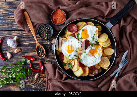 Petit déjeuner espagnol populaire d'oeufs au plat frit croustillant avec pommes de terre, saucisses de porc émincé dans une poêle sur une table en bois, cuisi Banque D'Images