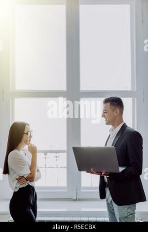 Heureux couple de jeunes entreprises partenaires qui travaillent dans un bureau moderne. Deux collègues travaillant sur un ordinateur portable en étant debout près de la fenêtre. Banque D'Images