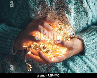 Coeur chaud, l'amour gratuit. Women's Hands holding a garland. Fille dans un pull bleu avec des lumières de Noël dans ses mains, chaude ambiance de Noël, soft focus. Banque D'Images