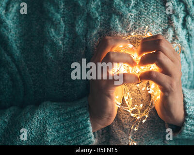 Coeur chaud, l'amour gratuit. Women's Hands holding a garland. Fille dans un pull bleu avec des lumières de Noël dans ses mains, chaude ambiance de Noël, soft focus. Banque D'Images