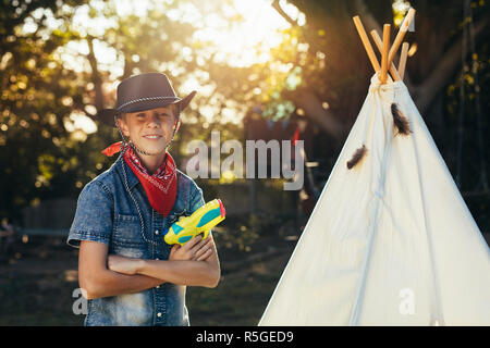 Petit cowboy debout à l'extérieur par un jouet hut en cour avec pistolet à eau. Young boy wearing cowboy hat squirt gun avec de l'eau. Banque D'Images