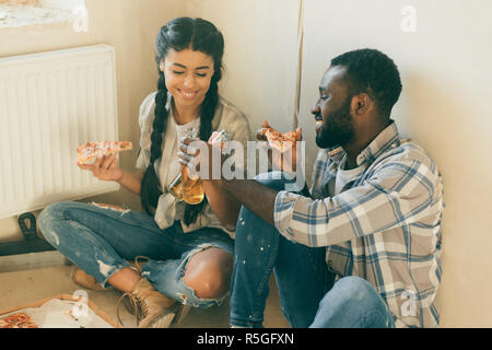 Happy young couple tintement des bouteilles de bière et de manger une pizza tout en effectuant la rénovation d'accueil Banque D'Images