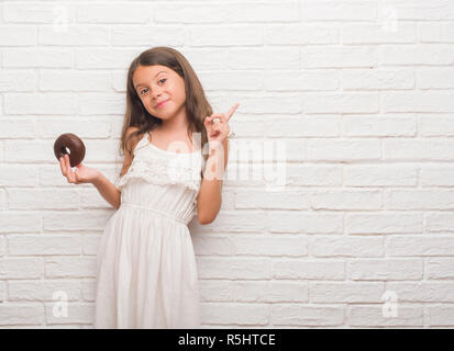 Les jeunes enfants hispaniques au mur en brique blanche manger du chocolat donut très heureux pointant avec la main et le doigt sur le côté Banque D'Images