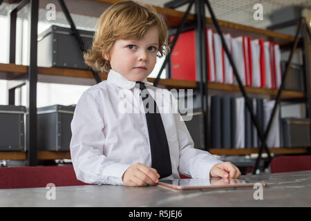 Petit garçon en chemise et cravate est debout dans le bureau et à l'aide de tablette. Banque D'Images