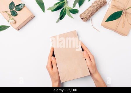 Woman's hands holding sac en papier brun sur fond blanc au milieu du cadre de mise à plat de feuilles vertes, de cadeaux et de fleurs rose, vue du dessus. Banque D'Images