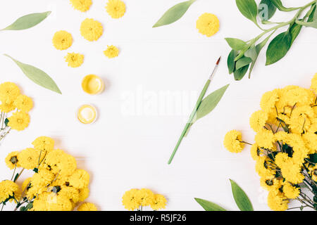 Carte de vœux vide à côté du pinceau et de la peinture à la gouache jaune printemps parmi les fleurs et feuilles vertes sur le tableau blanc, vue du dessus. Banque D'Images