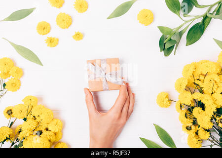 Vue de dessus de woman's hand holding petit cadeau fort parmi les fleurs jaune vif sur fond blanc, télévision lay. Banque D'Images