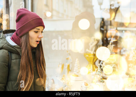 Une belle jeune femme ou de la fille ressemble à travers une vitrine lors d'une vacances de Noël et s'étonne. Banque D'Images