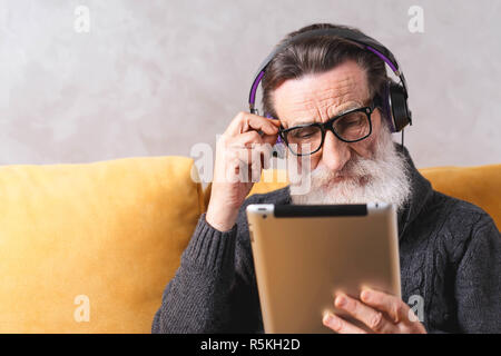Homme barbu graves supérieurs en portant des lunettes laser pull gris considérant la tablette numérique tout en restant assis dans le casque sur un canapé jaune lumière dans son salon. Banque D'Images