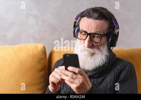 Homme barbu contemporain senior dans le port de lunettes pull gris message écrit sur le smartphone tout en restant assis dans le casque sur un canapé jaune dans sa lumière, salon de la technologie moderne, communication concept Banque D'Images