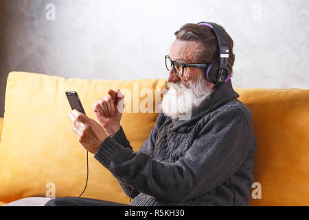 Joyeux Senior homme barbu portant des lunettes en pull gris smartphone à l'aide tout en restant assis dans le casque sur un canapé jaune dans sa lumière, salon de la technologie moderne, communication concept Banque D'Images