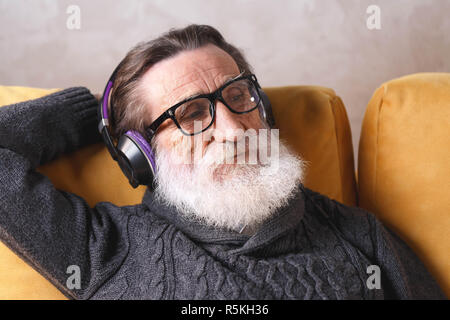 Beau Senior homme barbu portant des lunettes en pull gris assis dans le casque sur un canapé jaune dans sa lumière salle de séjour, de détente et d'écouter sa musique préférée, la technologie moderne, communication concept Banque D'Images