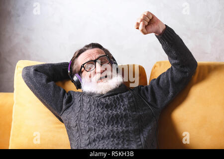 Beau Senior homme barbu portant des lunettes en pull gris assis dans le casque sur un canapé jaune dans sa lumière salle de séjour, de détente et d'écoute sa musique rock chanson, il fermer les yeux et inscrivez-vous à la chanson, la technologie moderne, communication concept Banque D'Images