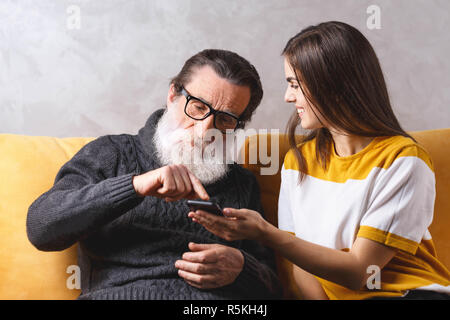 Fille brune aux cheveux long adultes explique à son père barbu senior dans les verres comment utiliser le smartphone tout en étant assis sur le canapé jaune à la lumière salle de séjour, la technologie moderne, communication concept Banque D'Images